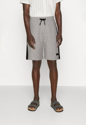 Widok z przodu osoby w szarych szortach ze ściągaczem i czarnymi paskami po bokach, białej koszuli w paski oraz szarych sandałach z klamrą.