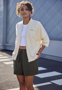 Hellgelbe Zip-Jacke mit weiten Ärmeln und Fronttaschen, kombiniert mit dunkelgrünen Shorts mit Kordelzugbund und Seitentaschen.