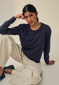 Maglione lungo navy a maniche, con colletto a coste e spalle a palloncino, abbinato a pantaloni a gamba larga color crema e ballerine turchesi. Sfondo neutro.