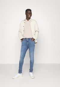 Chaqueta beige claro sobre una camiseta polo rosa de canalé; jeans ajustados azul; zapatillas blancas con suela texturada; fondo neutro.