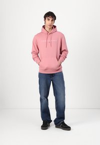 Felpa rosa con logo, misto cotone, tasca frontale, coulisse regolabile. Abbinata a jeans blu e sneakers nere. Fit semplice e casual.