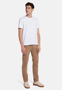 T-shirt bianca in cotone a maniche corte, abbinata a pantaloni slim-fit marrone chiaro e sneakers beige con suola marrone. Design minimalista.