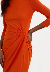 Vestido laranja justo ao corpo com mangas longas, apresentando um detalhe atado na cintura e tecido drapeado para uma silhueta elegante.