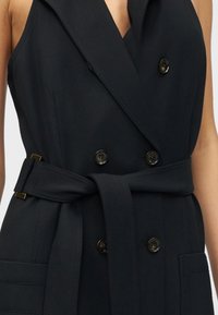 Μαύρο αμάνικο διπλόστενο γιλέκο με μεγάλα κουμπιά, φαρδιά ζώνη δεμένη στη μέση και τσέπες με καπάκι, φορεμένο στον κορμό ενός ατόμου.