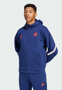 Sudadera con cremallera en azul marino con un logo de gallo rojo y rayas blancas en las mangas. Hecha de tela suave, con un bolsillo frontal y capucha.