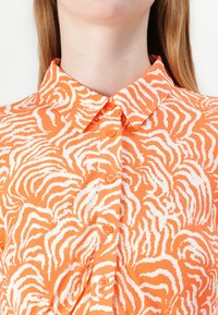 Oranssi nappikauluspaita, jossa on valkoisen tiikeriraidan kuvio, klassinen kaulus ja teksturoitu kangas. Etupainikkeet näkyvät.