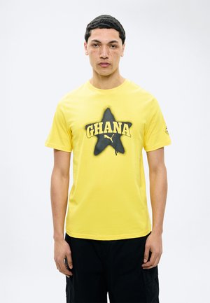 Jeune homme portant un T-shirt jaune avec une étoile noire et le texte "GHANA", assorti à un pantalon noir, debout devant un fond blanc uni.