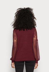 Bordeauxrode trui met lange doorschijnende mouwen met bloemmotieven, geribbelde manchetten en zoom, en een aansluitend lijfje.