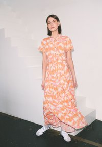 Maxi sukienka z krótkimi rękawami, pomarańczowy i biały wzór tie-dye, zapięcie na guziki z przodu, ściągnięta w talii, teksturowany materiał, zestawiona z białymi sneakersami.
