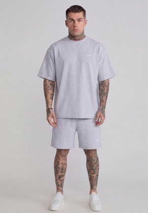 Mužský model s tetovaniami na rukách a nohách, oblečený v šedom tričku SikSilk a zodpovedajúcich šortkách s bielymi teniskami, stojaci pred jednofarebným pozadím.