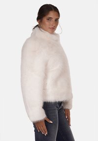 Giacca in pelliccia sintetica bianca con colletto alto, vestibilità ampia e superficie texturizzata. Indossata sopra jeans scuri, con maniche lunghe e larghe.