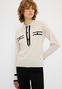 Polo a maglia a maniche lunghe crema con accenti e bottoni neri, caratterizzato da un colletto a punta e due tasche sul petto con dettagli in nero.