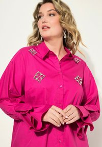 Fuchsia Hemdbluse mit verzierten Schultern, die mit dekorativen Kristallen ausgestattet ist. Lange Ärmel mit gerafften Manschetten und glatter Textur.