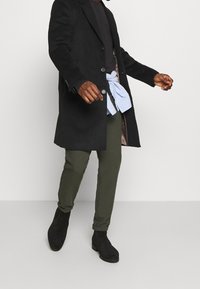 Hombre con abrigo negro, suéter oscuro, camisa azul atada a la cintura, pantalones oliva y botas negras hasta los tobillos, de pie contra un fondo claro y liso.