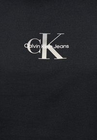 Λογότυπο Calvin Klein Jeans με μεγάλα γκρι γράμματα "cK" και μικρότερο λευκό κείμενο "Calvin Klein Jeans" σε μαύρο υφαντό φόντο.