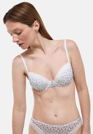 Femme aux cheveux bruns et lisses, portant un soutien-gorge blanc orné de dentelle fleurie et une lingerie assortie, regardant vers le bas à sa gauche sur fond blanc.