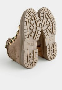 Stivali in suede beige con suola in gomma testurizzata e un motivo di battistrada robusto, arricchiti da dettagli dorati lungo i lacci, che migliorano la presa e lo stile.