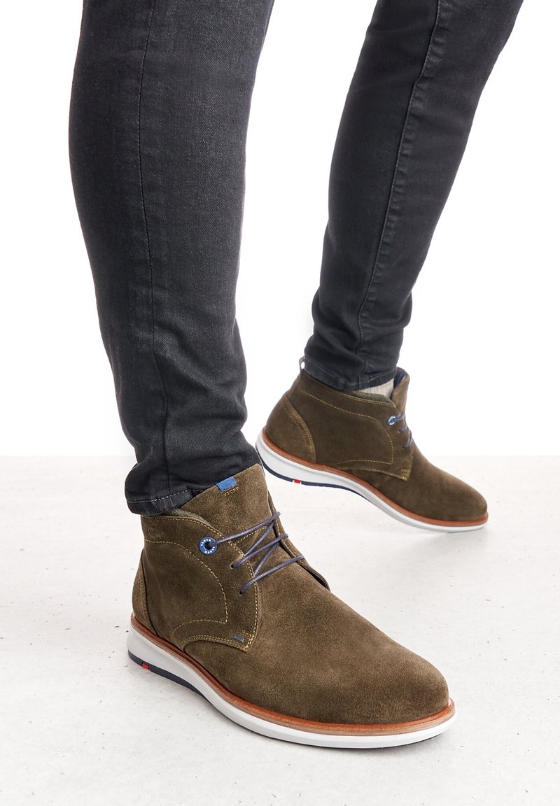 Personne portant un pantalon slim foncé et des bottines marron en daim à lacets avec semelles blanches, debout sur une surface claire.