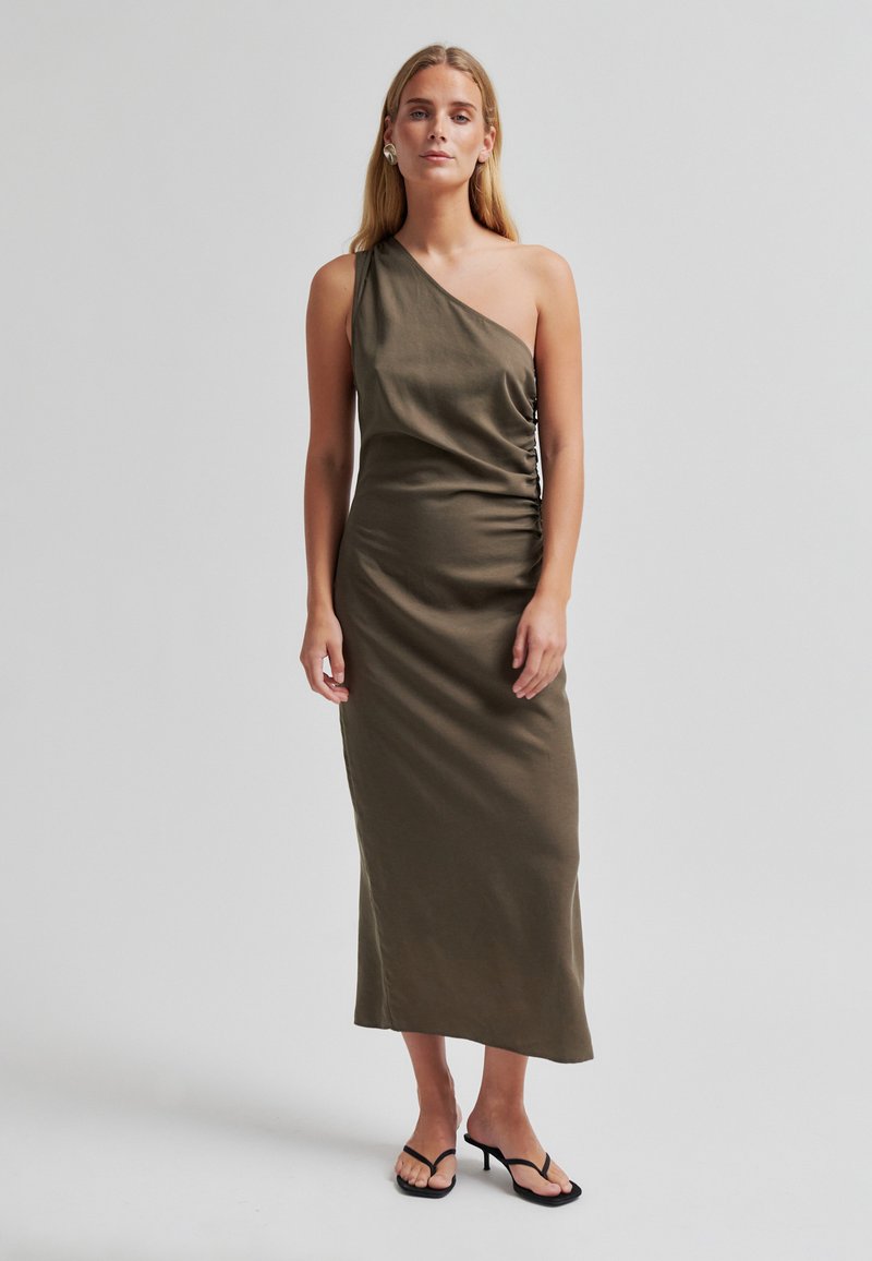 Femme portant une robe midi asymétrique vert olive avec un détail froncé sur le côté et des sandales à talons noires, debout devant un fond gris uni.