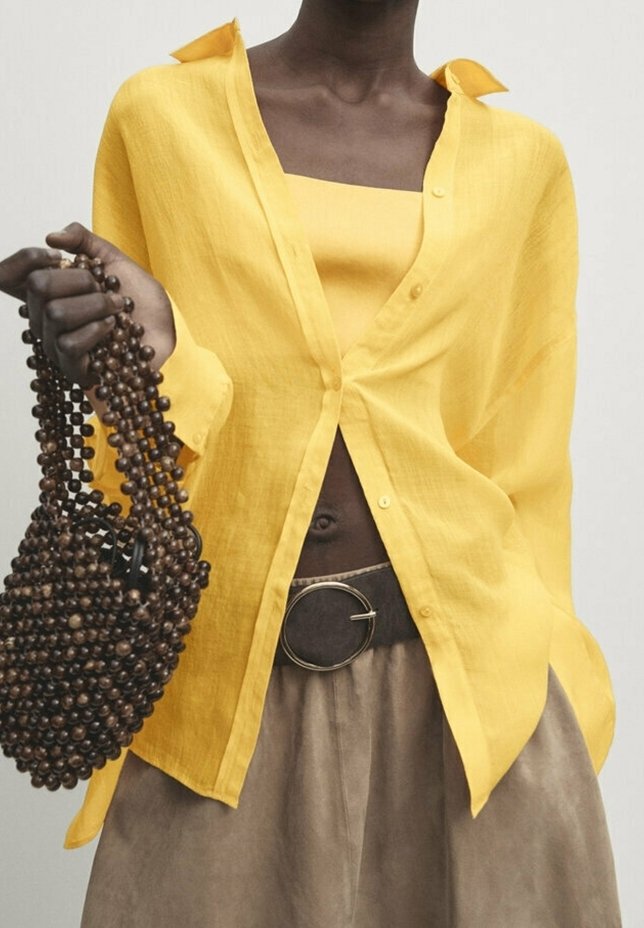 Personne portant une chemise jaune boutonnée et un haut, un pantalon beige avec une ceinture à boucle circulaire, tenant un sac à main marron orné de perles.