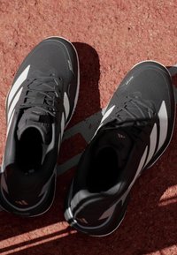 Czarne buty sportowe z teksturowaną cholewką, z białymi paskami i amortyzowaną podeszwą, umieszczone na czerwonej powierzchni.