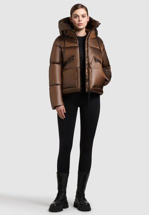 Brun puffer jakke med en glat finish, oversized hætte, to lynlåslommer foran og elastiske manchetter, båret over sorte leggings og kampstøvler.