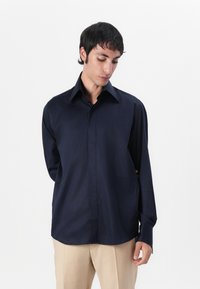Cămașă lungă de culoare bleumarin, cu un guler clasic, textură fină, buzunar unic și tiv dreaptă, purtată cu pantaloni bej deschis.