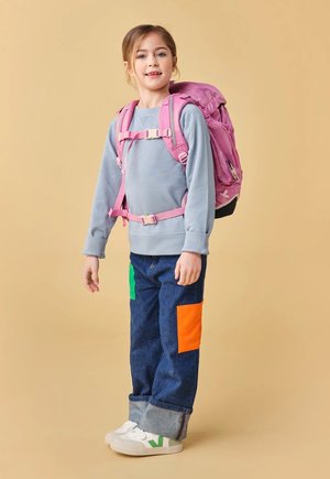 Junges Mädchen trägt einen grauen Pullover, Jeans mit Flickenelementen, weiße Turnschuhe und einen rosa Rucksack mit Brust- und Taillengurten vor beigem Hintergrund.