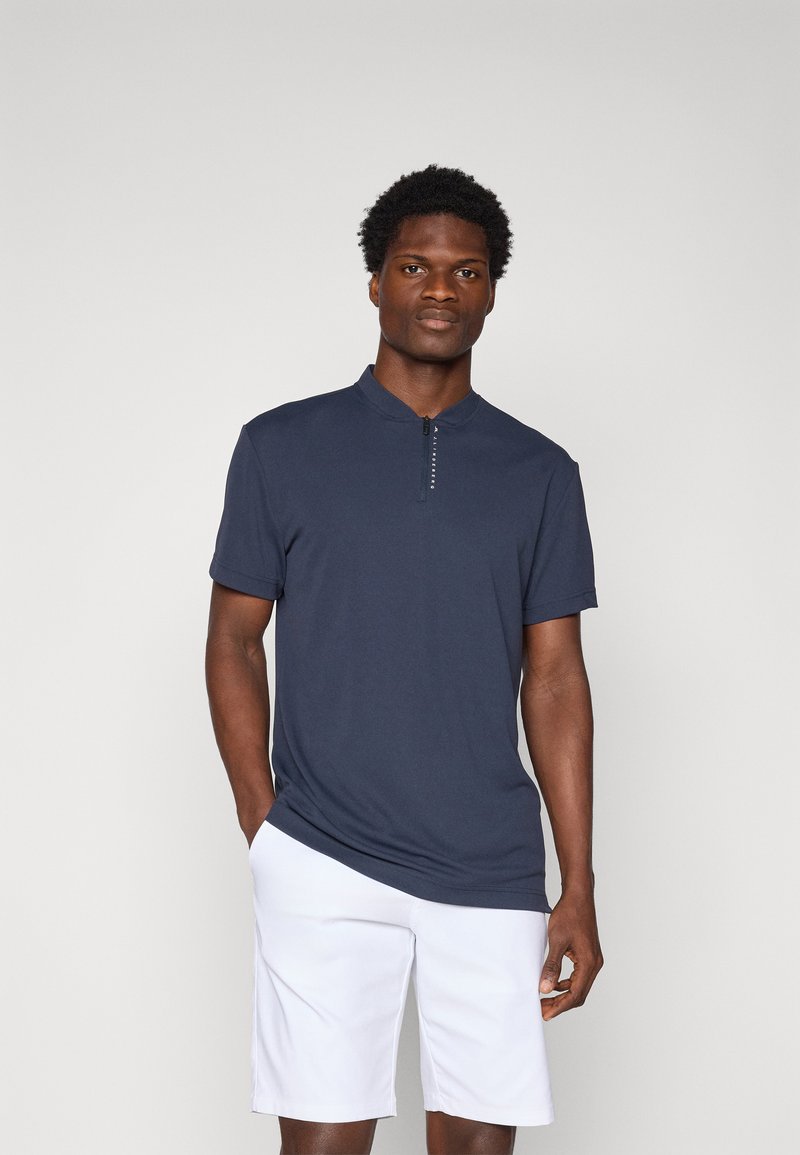 Polo din bumbac navy cu mânecă scurtă, având guler în picioare, cu un fermoar frontal și o textură netedă. Poartă o pereche de pantaloni scurți albi.