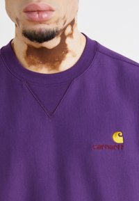 Violetinis džemperis su grioveliais apykaklėje, ant kairiojo krūtinės pločio pasiūta maža geltona ir raudona Carhartt logotipo siuvinėjimas.