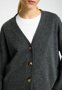 Šedý kardigan z vlněné směsi s hlubokým výstřihem do V, předním zapínáním na knoflíky s hnědými knoflíky a ribovanými manžetami a lemem.