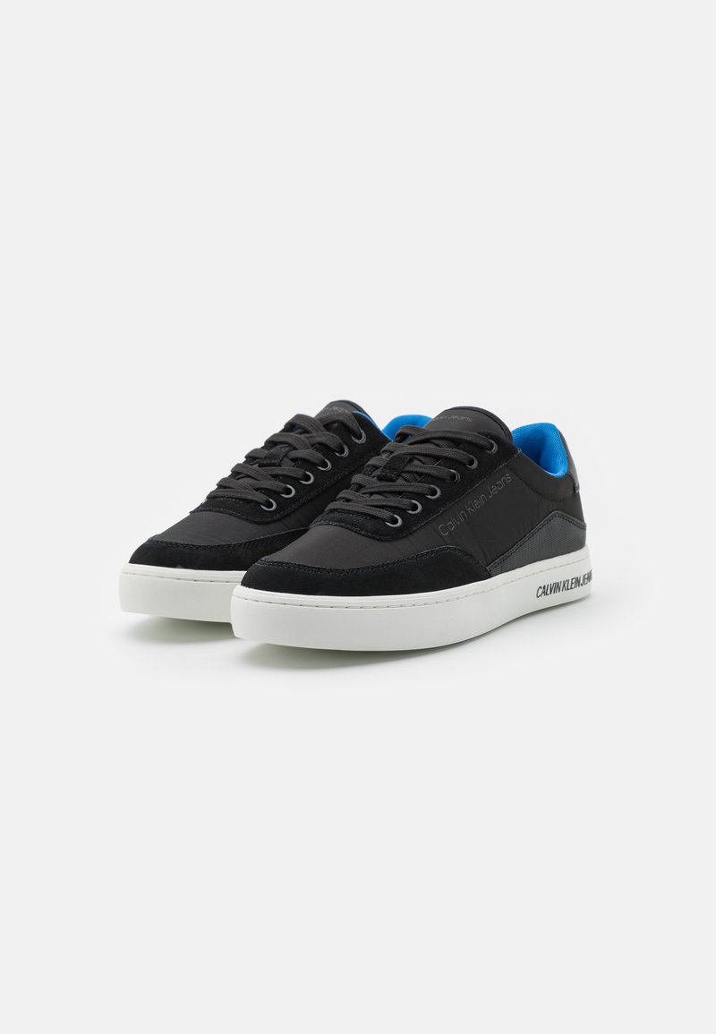 Baskets en daim noir avec semelle en caoutchouc blanche, présentant une doublure bleue, un bout rond et des lacets plats. Marque sur le côté.
