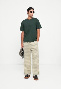 Tricou verde din bumbac cu logo, pantaloni largi bej cu carouri, saboti negri cu talpă maro și ochelari de soare, ținând o sapcă gri.