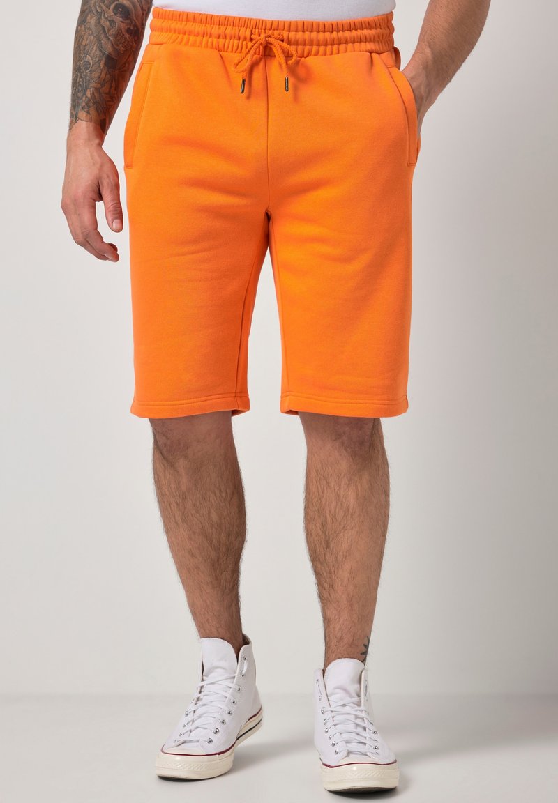 Mann trägt leuchtend orangefarbene Shorts mit Kordelzug und weiße High-Top-Sneaker, steht mit teilweise in den Taschen steckenden Händen.