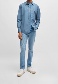 Camisa de botones de color azul claro con mangas largas, combinada con vaqueros azul claro y zapatillas blancas con acentos en beige. Tejido de algodón suave.