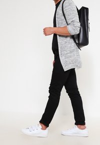 Grå stickad cardigan över en svart skjorta, kombinerad med svarta byxor och vita sneakers. Ryggsäck är svart med en enkel design.