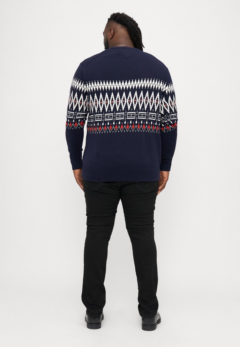 Navy knitted sweater with a geometric white, red, and blue pattern across the shoulders; paired with black pants and shoes.