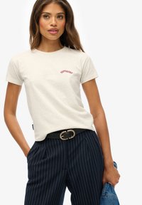 Kobieta w beżowym t-shircie Superdry, granatowych spodniach w prążki i czarnym pasek, trzyma w jednej ręce dżinsową kurtkę.