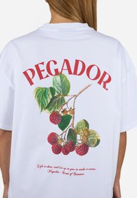 Fehér pamut póló, színes málna és levél grafikával. A "PEGADOR." felirat vörös színben, hangsúlyosan szerepel a minta felett.