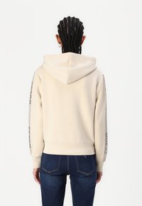 Sudadera beige con capucha, puños y dobladillo acanalados. Presenta texto oscuro a lo largo de las mangas. Se usa con jeans ajustados de mezclilla oscura.