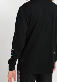 Svart långärmad sporttröja i meshmaterial, med perforerade detaljer och färgglada ränder på vänster ärm.