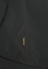 Gros plan sur un tissu noir avec une petite étiquette noire comportant le texte jaune "TEXAPORE" cousue près d'une couture.