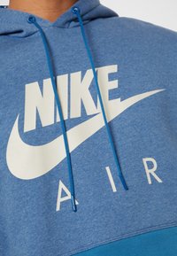 Blå fleece hoodie med en stor vit Nike-logotyp och texten "AIR". Inkluderar blå dragkedjor och en texturerad tygfinish.
