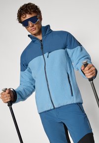 Giacca in pile blu con una sezione superiore a contrasto; presenta una tasca con zip e un tessuto testurizzato, abbinata a pantaloni blu coordinati e bastoncini da trekking.