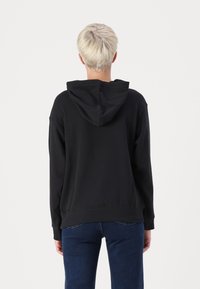 Felpa nera con vestibilità rilassata, polsini a costine e un grande cappuccio. Il tessuto sembra morbido e leggermente strutturato. Indossata con jeans blu scuri.