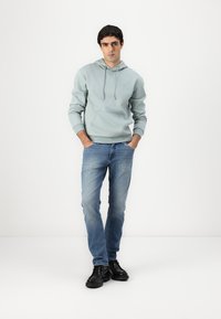 Blusão azul claro com cordões e bolso frontal, combinado com jeans azul claro e botas pretas de tornozelo, em pé contra um fundo branco.