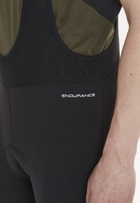Svarta cykelbyxor med hängslen och en grön och svart överdel. Har texten "ENDURANCE" i vitt. Mjukt, elastiskt tyg för komfort.