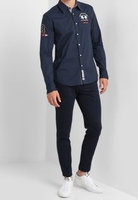 Camisa de botones de manga larga color azul marino con cuello, logo bordado en el pecho y un detalle numérico en la manga, combinada con pantalones oscuros y zapatillas blancas.
