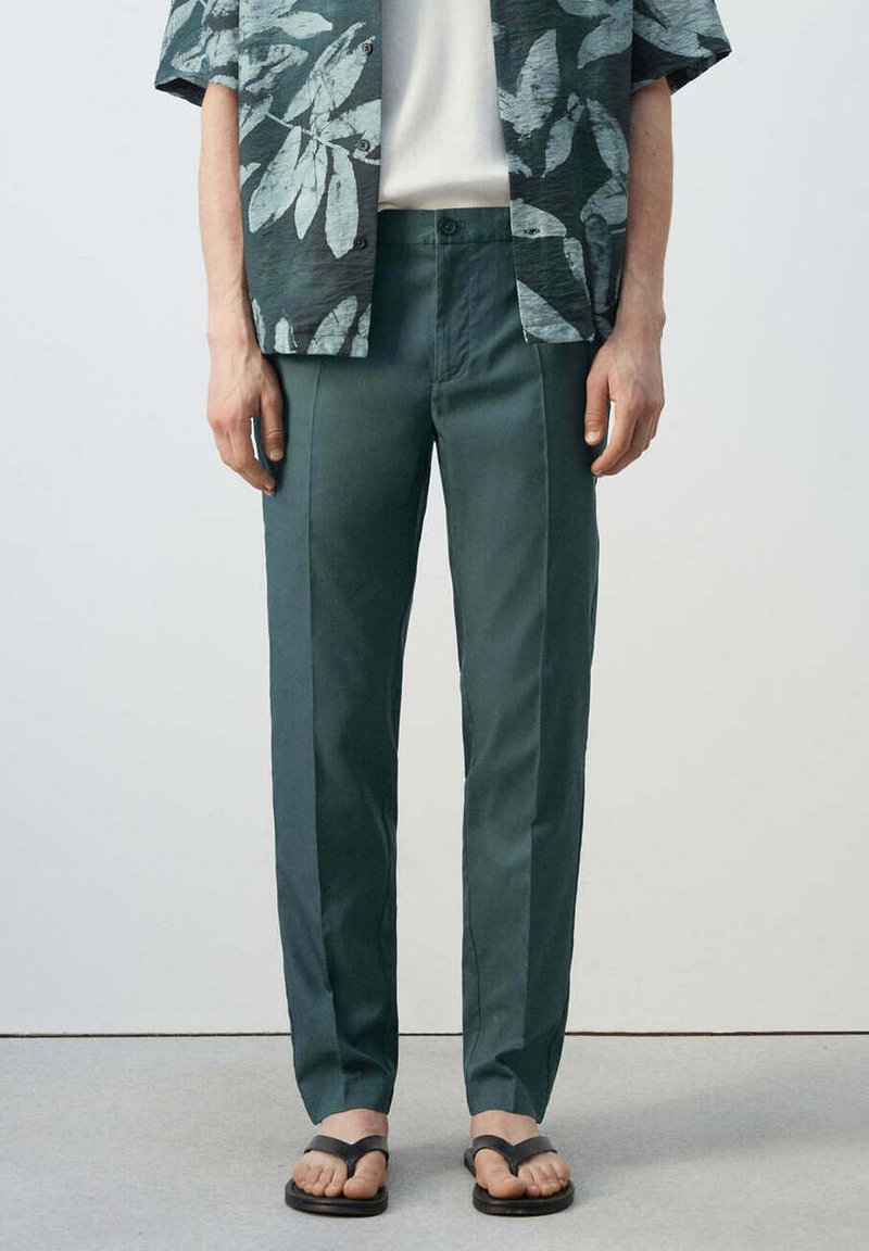 Homme portant un pantalon droit vert foncé, une chemise blanche, une chemise à manches courtes fleurie vert foncé et des sandales noires, debout.