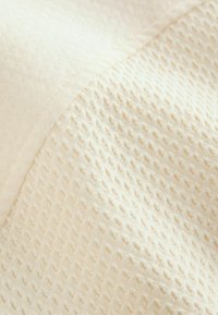 Beige texturerat tyg som visar två mönster: ett slätt och ett med små upphöjda rutor. Mjukt, lätt material med en lätt lyster.